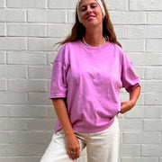 Person wearing a pink t-shirt and white pants standing against a white brick wall.
